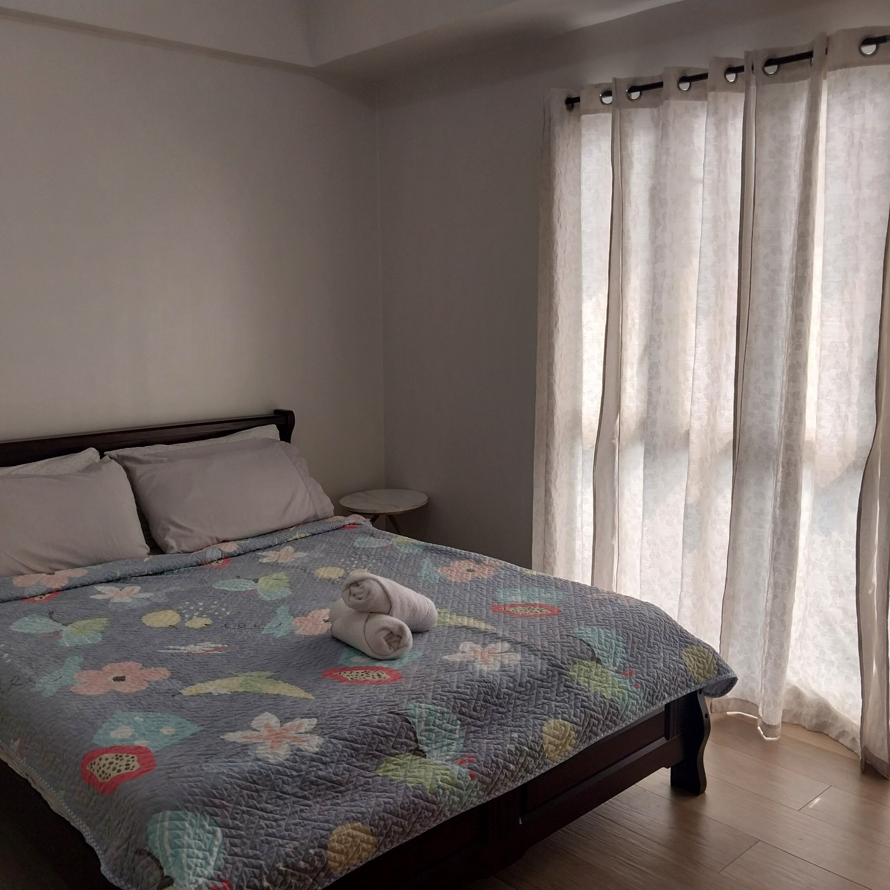 A neatly made bed with a floral quilt, a small bedside table, and a window with sheer curtains.