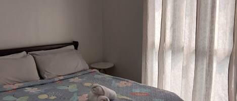 A neatly made bed with a floral quilt, a small bedside table, and a window with sheer curtains.