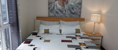 Habitación de hotel con una cama que tiene una colcha de estampado geométrico, un cabecero de madera y un cuadro de una persona en el agua en la pared.