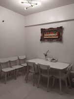 Een eethoek met een witte tafel en stoelen, een lijstwerk met een kunstwerk erboven en een plafondlamp.