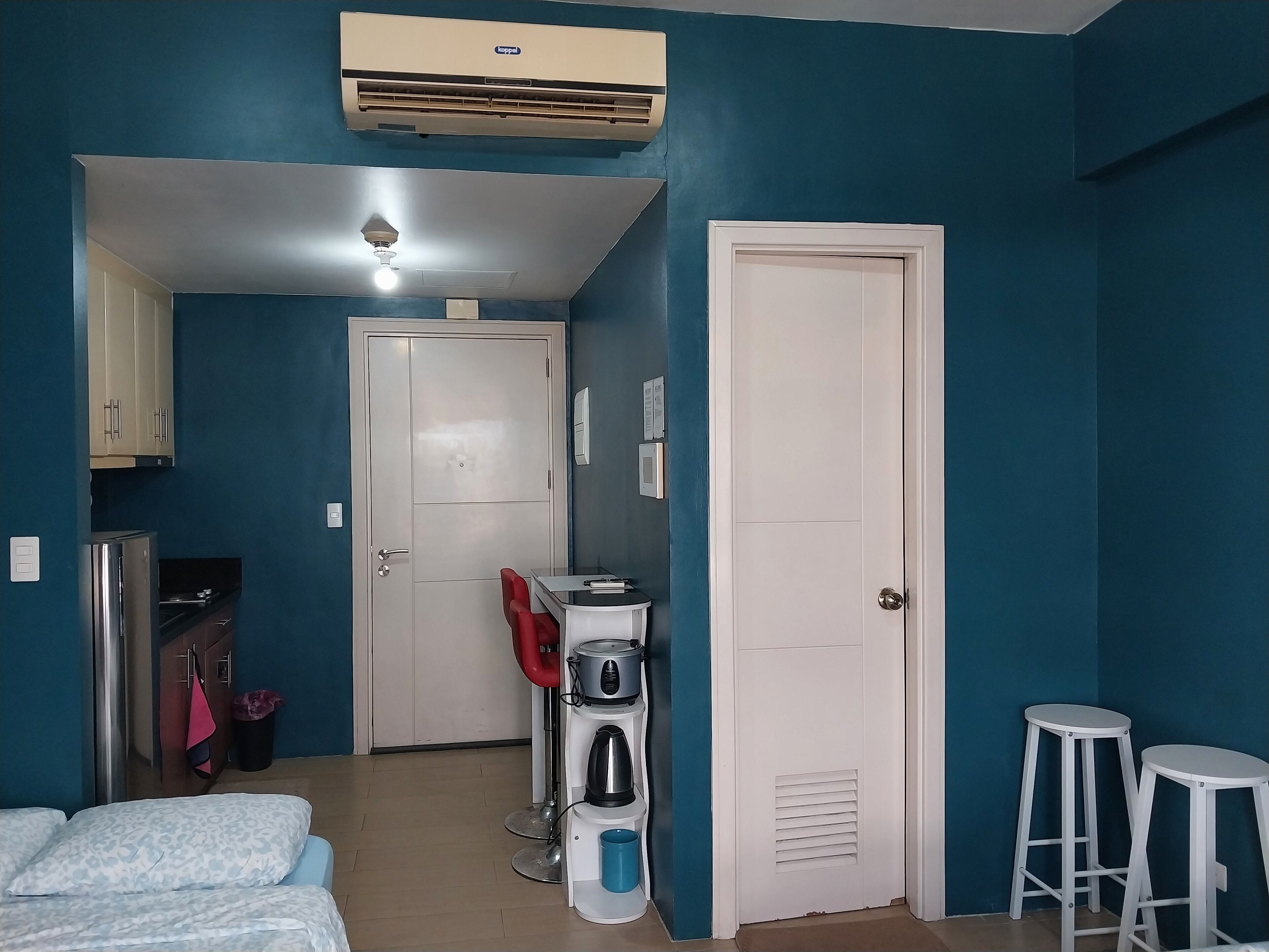 Ein Zimmer mit Türkiswänden, einer weißen Tür, einer Küchenzeile mit Kühlschrank, Herd und Spüle. Zwei weiße Hocker und ein Bett mit gemusterten Kissen.