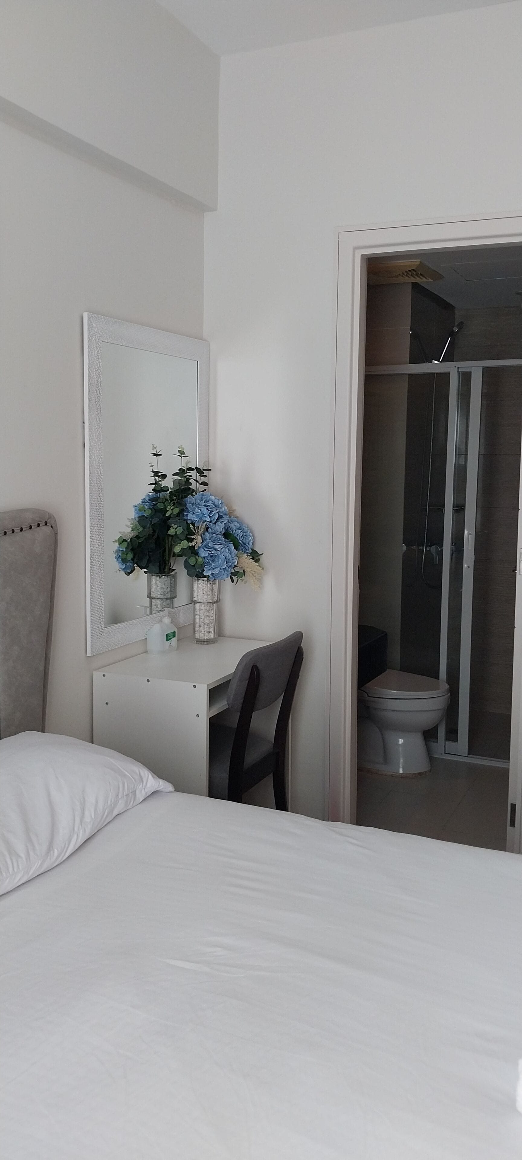 Un dormitorio con cama, escritorio y silla, espejo, y baño con inodoro y ducha.