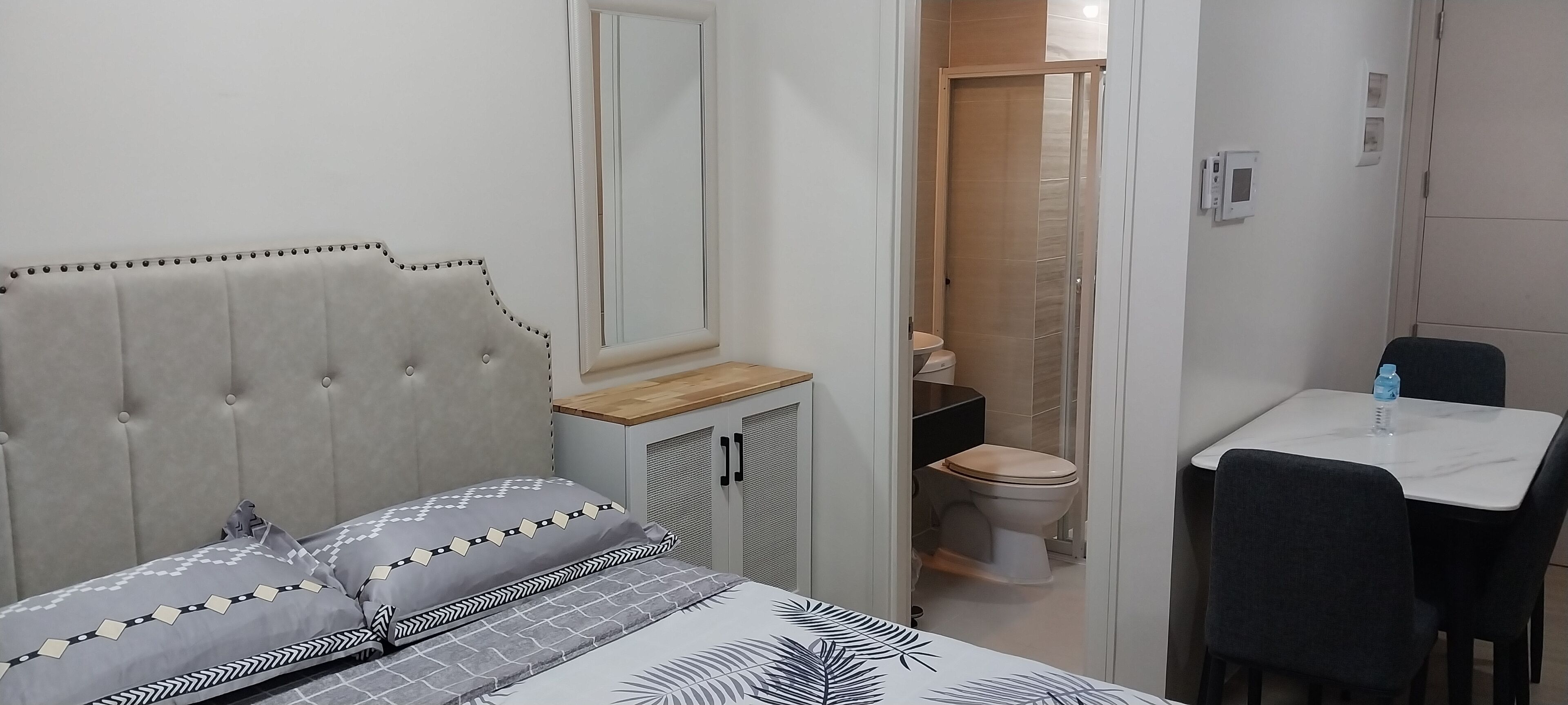 A bedroom with a bed, a bedside table, a chair, a small table with a water bottle, and a bathroom visible through an open door.