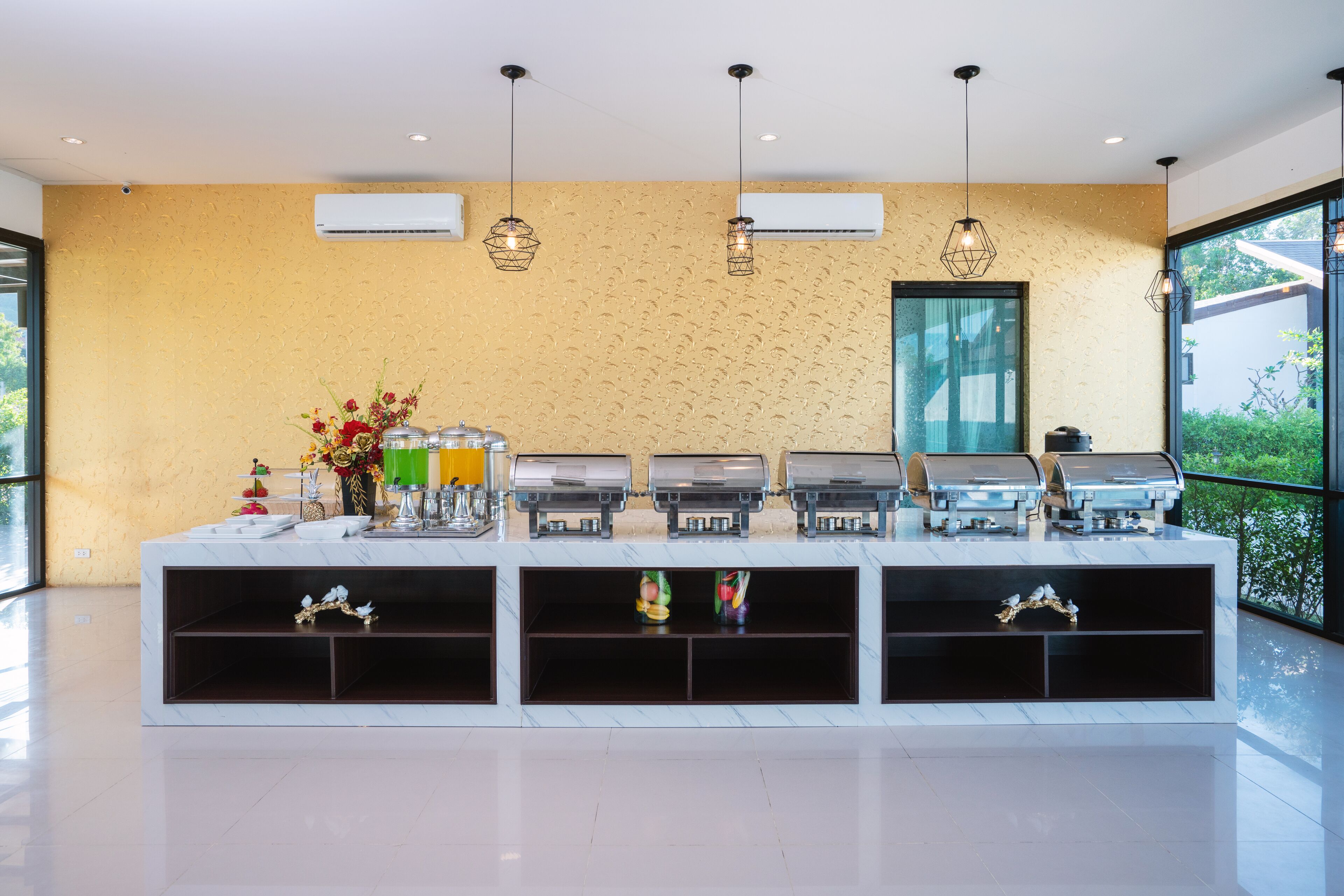 A buffet setup with multiple food dispensers, decorative vases, and hanging pendant lights in a modern room with a large window.