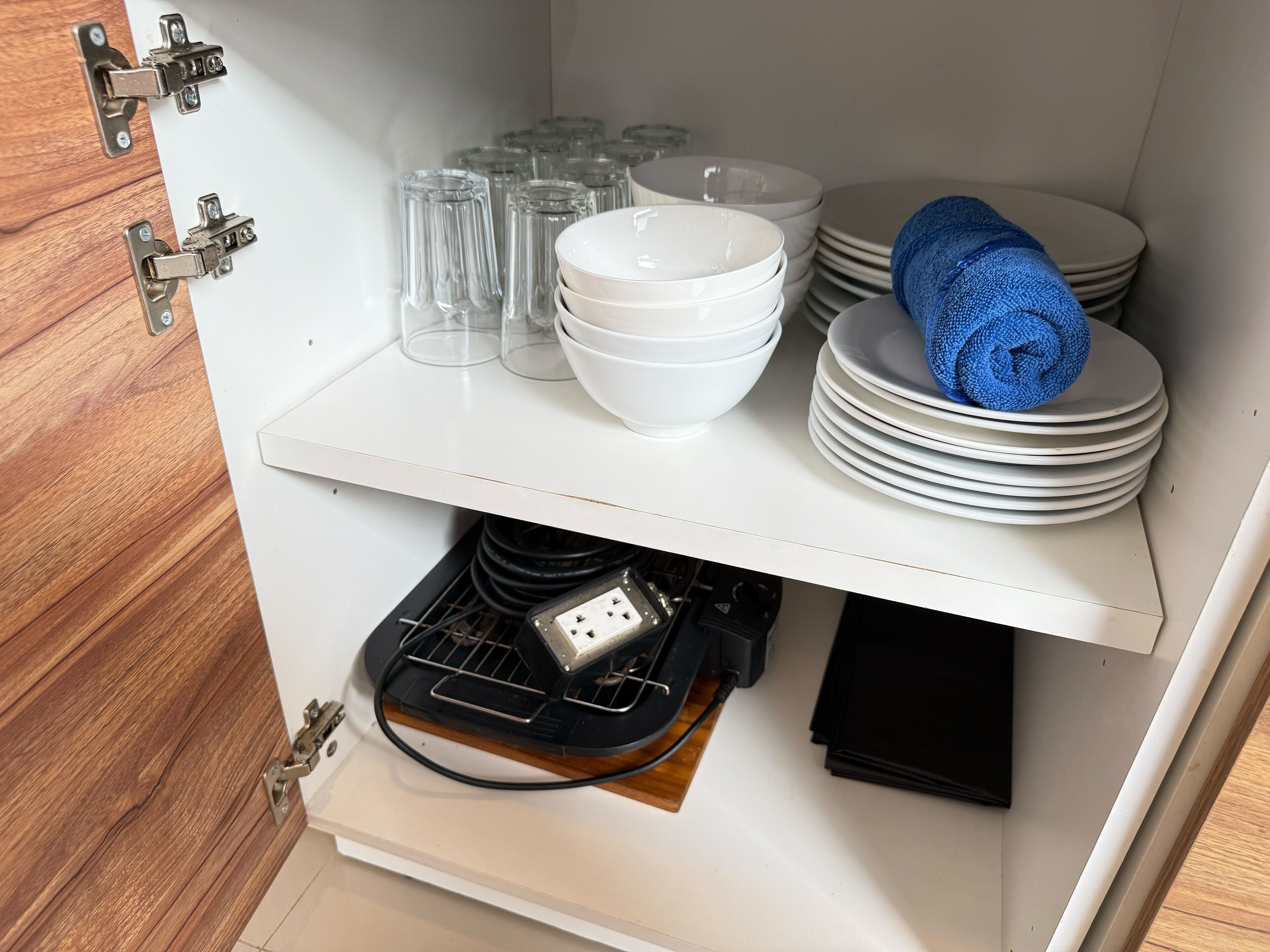 A kitchen cabinet with white plates, bowls, and glasses stacked on the upper shelf, and a black appliance on the lower shelf.