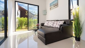 A modern living room with a sofa, a small table with a vase, and a large window offering a view of greenery and a building. - Thames Tara Private Pool Villa Rawai Phuket (Rawai)