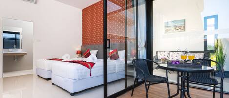 A modern hotel room with a large bed, a glass wall, a balcony with a table and chairs, and a small kitchen area.