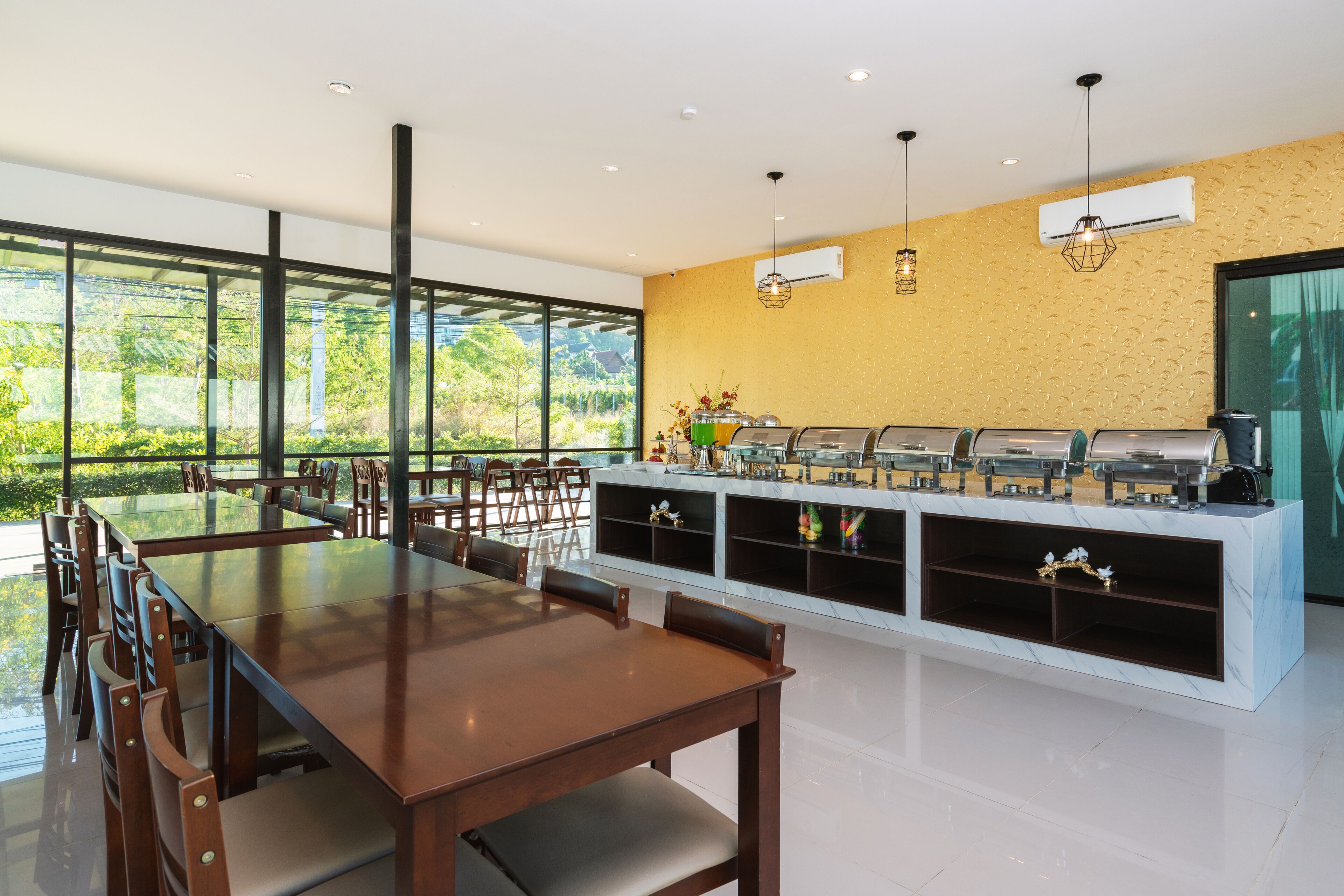 A modern dining area with a long wooden table and chairs, a buffet station with various food items, and large windows offering a view of greenery.