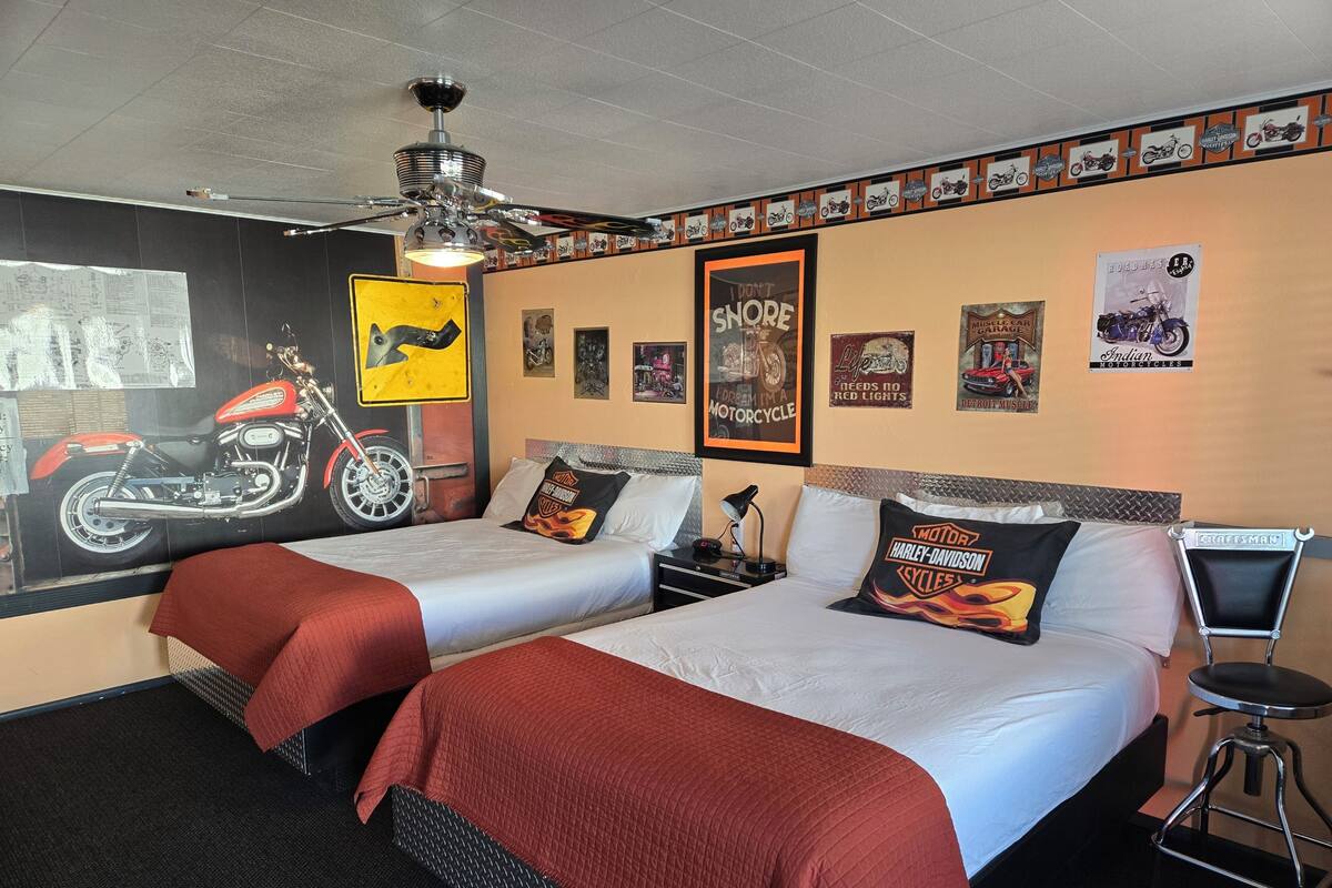 Habitación de hotel con dos camas, ventilador de techo, carteles en las paredes y decoración con temática de motocicletas.