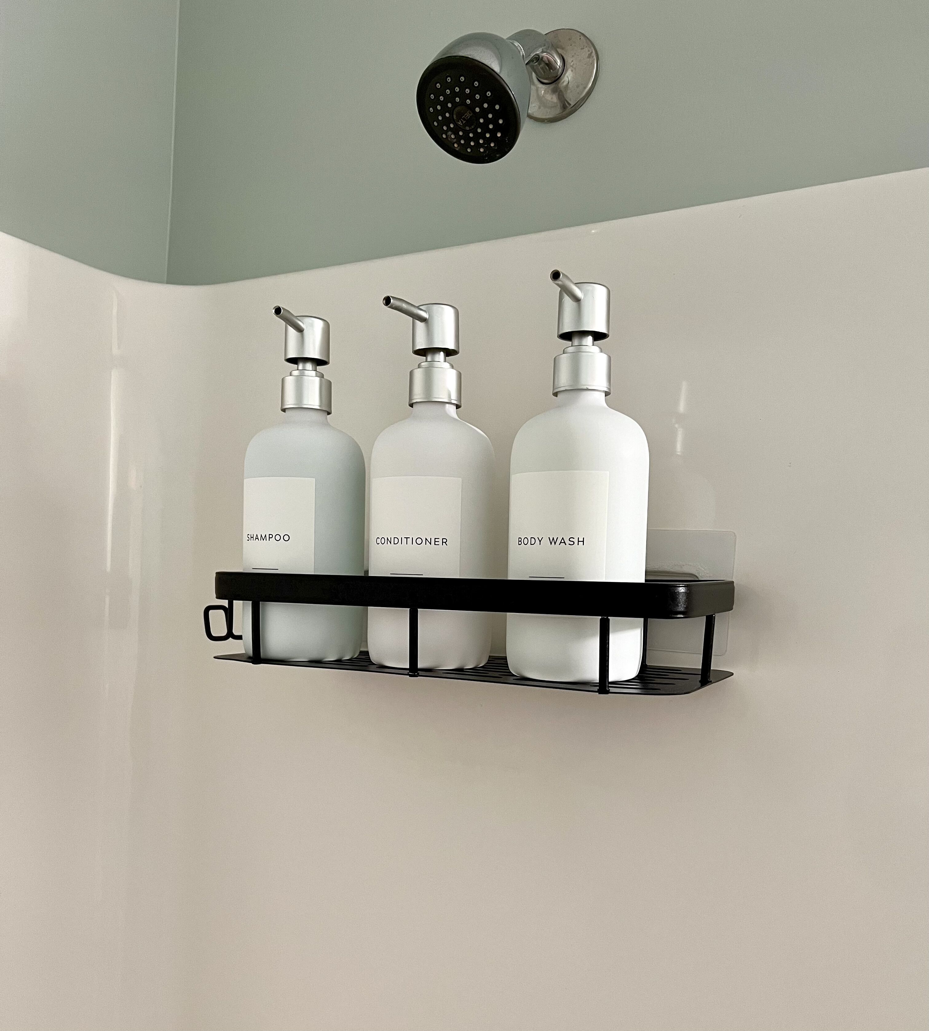 Baño con ducha, tres botellas de champú, acondicionador y gel de baño sobre un estante negro.