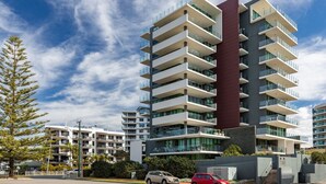 Un immeuble résidentiel moderne à plusieurs étages, doté de balcons, entouré d’autres bâtiments et de verdure.