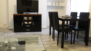 Un salon moderne avec un téléviseur à écran plat posé sur un meuble en bois, une table basse en verre et un coin repas avec une table et des chaises noires.