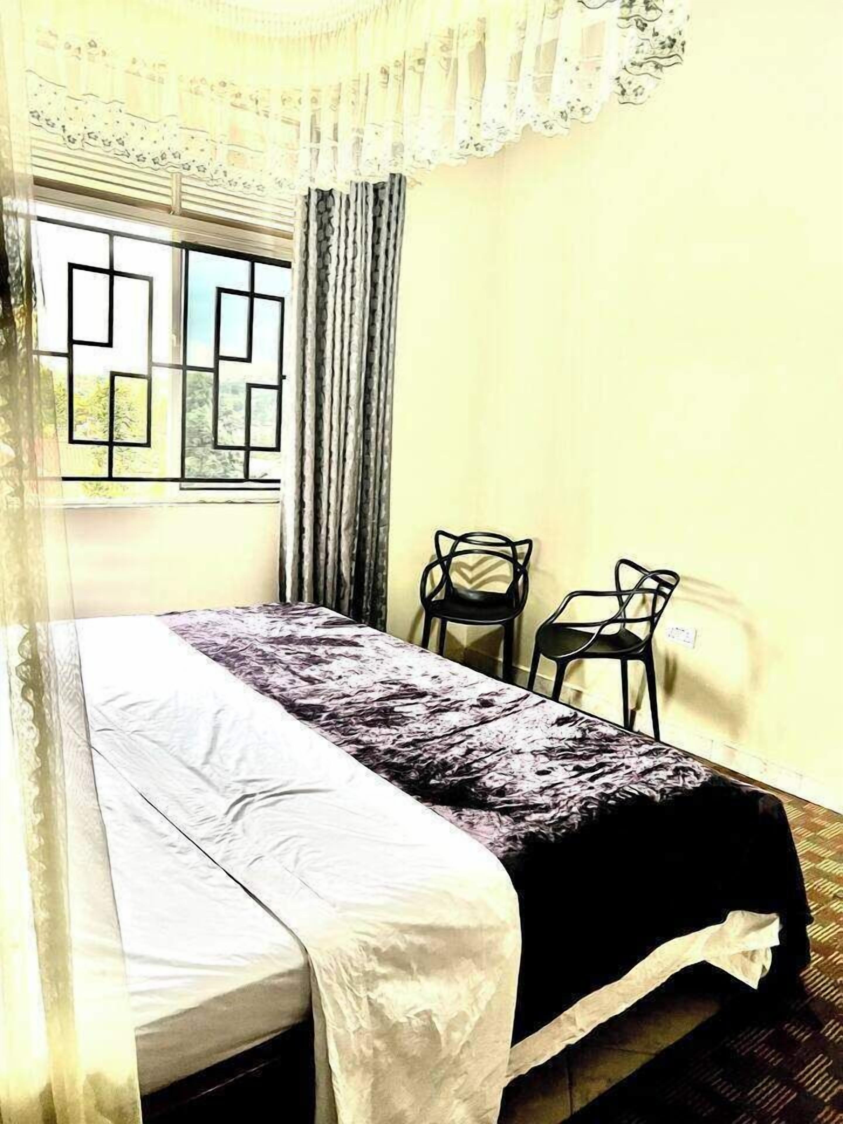 A room with a bed, two chairs, a window with a geometric pattern, and a curtain.