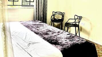 A room with a bed, two chairs, a window with a geometric pattern, and a curtain.
