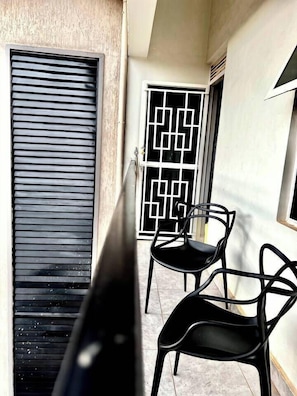 A balcony with two chairs, a window with a geometric pattern, and a door with horizontal slats.