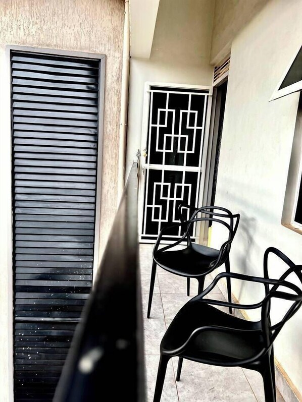 Un balcon avec deux chaises, une fenêtre ornée d’un motif géométrique et une porte à lames horizontales.