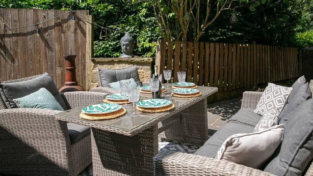 Eine Terrassesituation im Freien mit Korbmöbeln, einem Tisch, der mit Tellern und Gläsern gedeckt ist, und einer Buddha-Statue im Hintergrund.