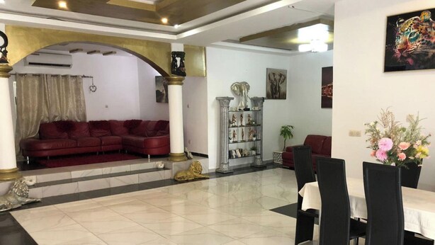 Amplia sala de estar con sofá rojo, una mesa de centro blanca y una mesa de comedor con sillas. Se observa un pilar decorativo y una estantería con figuritas.