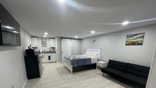 Un dormitorio con cama, un sofá negro, una mesita de noche, un tocador y una zona de cocina al fondo.