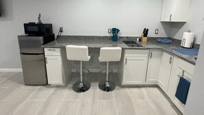 Cocina compacta con refrigerador, microondas, dos taburetes tipo barra, un lavamanos y una encimera con diversos objetos.