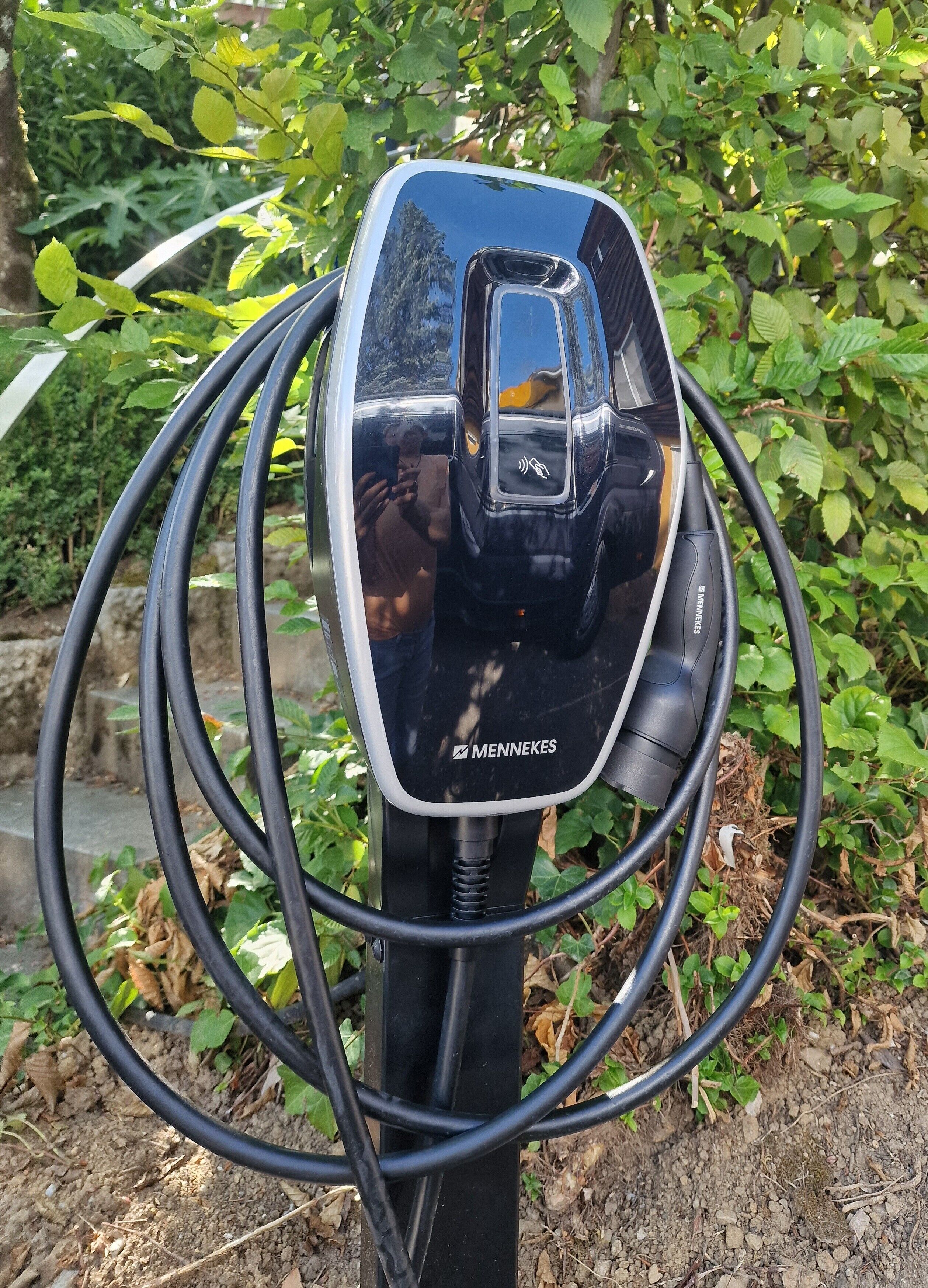 A black electric vehicle charging station with a visible brand logo, surrounded by greenery.