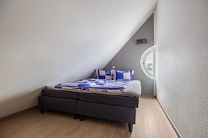 A bedroom with a sloped ceiling, a bed with a blue and white patterned bedspread, a round window, and a small wall-mounted light.