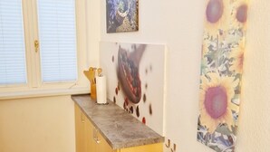Una cocina con encimera de madera, un fregadero y un cubo de basura azul. Hay dos ventanas con persianas, un cuadro en la pared y un horno empotrado.