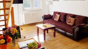 Un salón con escalera de madera, un sofá, una mesa de comedor con fruta, un televisor y una ventana con persianas.