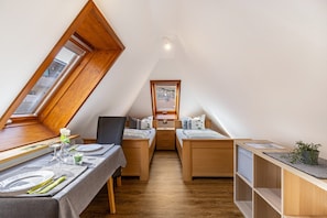 Habitación pequeña con techo inclinado, una mesa de comedor con sillas, dos camas individuales, un ventanal y una estantería con una planta.