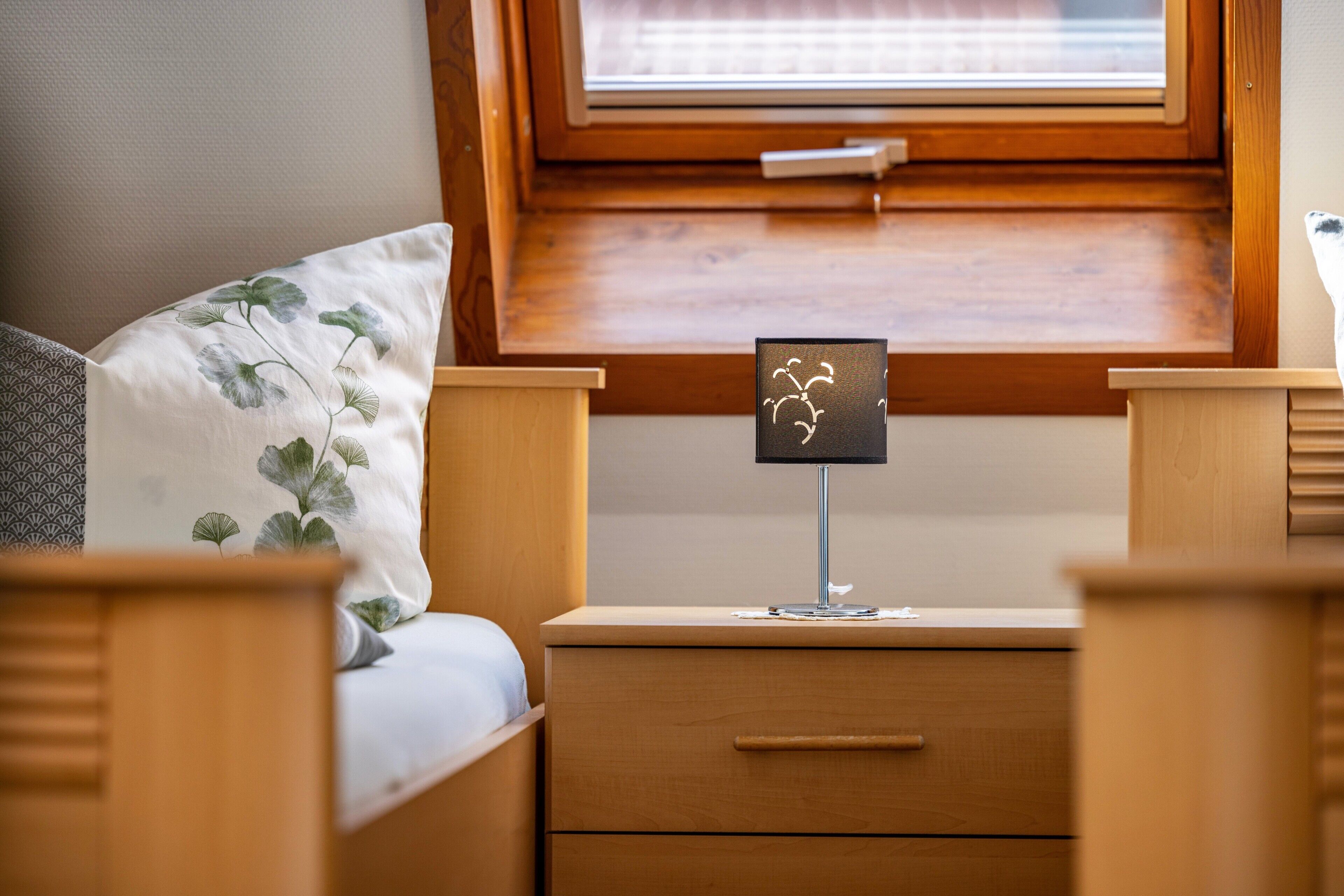 Een slaapkamer met een bed, nachtkastje met lamp, raam met jaloezieën en een houten hoofdbord.