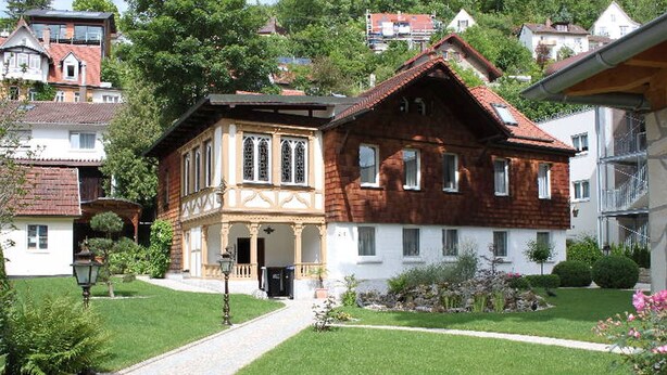 A traditional building with a central entrance, a garden with a paved path, and surrounding residential buildings. - Apartment 'Güterstein' with Mountain View, Shared Terrace and Wi-Fi (Bad Urach)
