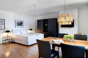 Une chambre d’hôtel moderne avec un lit, une table à manger en bois, des chaises noires, un meuble noir et un luminaire suspendu.