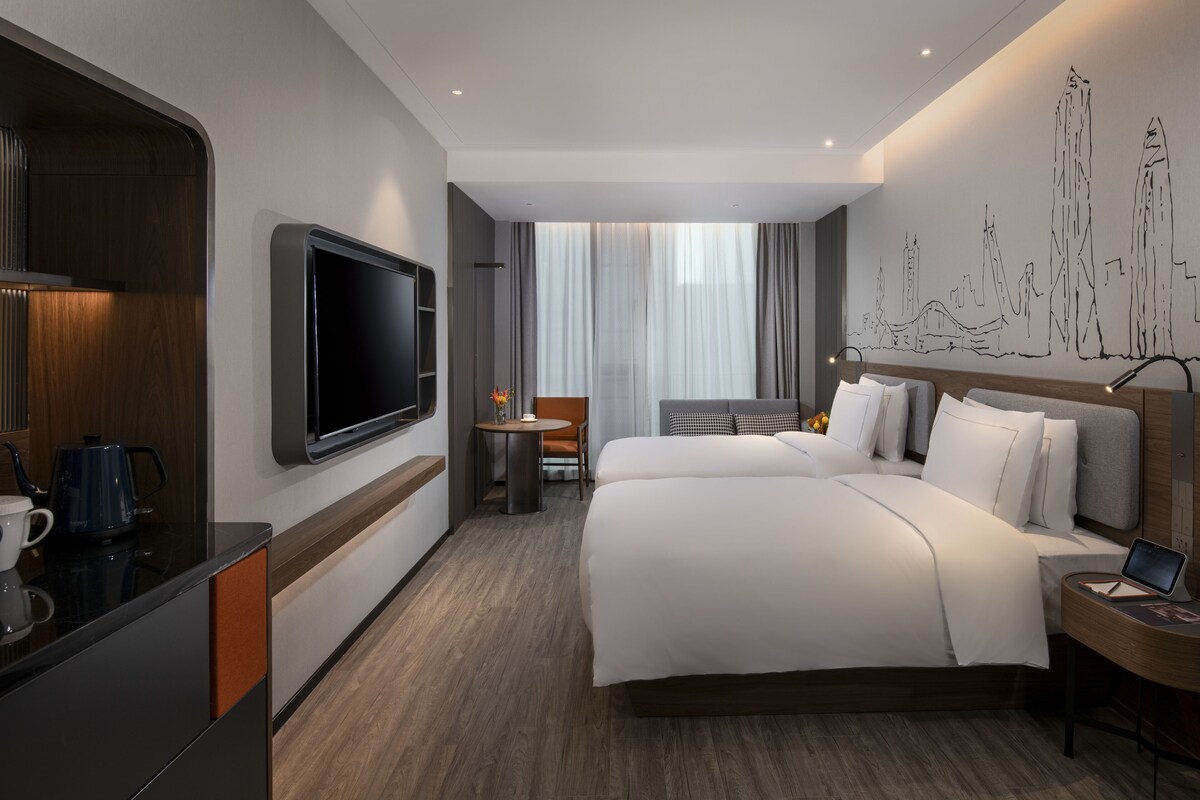 Une chambre d’hôtel moderne dotée d’un grand lit, d’un téléviseur à écran plat, d’un bureau avec un ordinateur portable et d’une fresque murale représentant un paysage urbain.