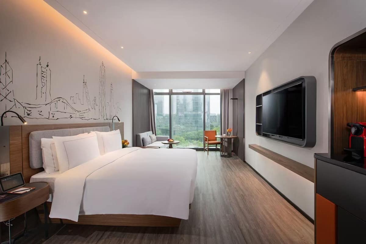 A modern hotel room with a large bed, a desk, a flat-screen TV, and a city view.