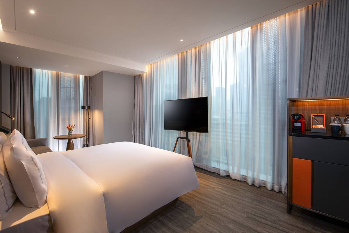 A modern hotel room with a large bed, a TV on a stand, a small table with a vase, and a minibar.