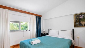 Ein Zimmer mit einem Bett, einem Fenster, Vorhängen, einem Nachttisch und einem Wandbild.