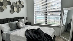 Een slaapkamer met een bed, een nachtkastje, een lamp, een groot raam met jaloezieën en een spiegel.