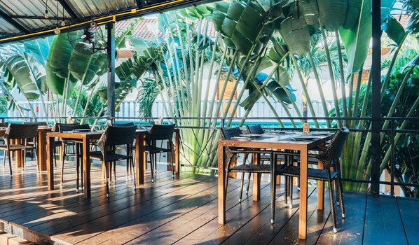 Un espacio exterior con mesas y sillas de madera, rodeado de hojas de plátano y plantas tropicales.