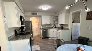 Cocina compacta con armarios blancos, encimera de granito y comedor con mesa para dos.