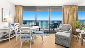 Un balcon avec vue sur la mer, une chaise blanche avec un coussin à motifs, un canapé gris et une petite table avec une lampe.