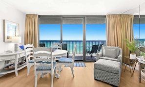 A balcony with a view of the sea, a white chair with a patterned cushion, a grey sofa, and a small table with a lamp.
