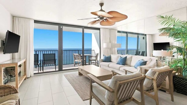 A spacious living room with a large sofa, wooden chairs, a coffee table, and a ceiling fan. A balcony with a glass door leads to a terrace with a view of the sea. - One Seagrove Place 1503 (Santa Rosa Beach)