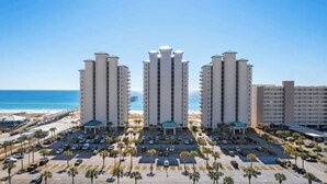 Ein Hotel mit drei hohen Gebäuden, einem Parkplatz mit Autos und einem Strand im Hintergrund.