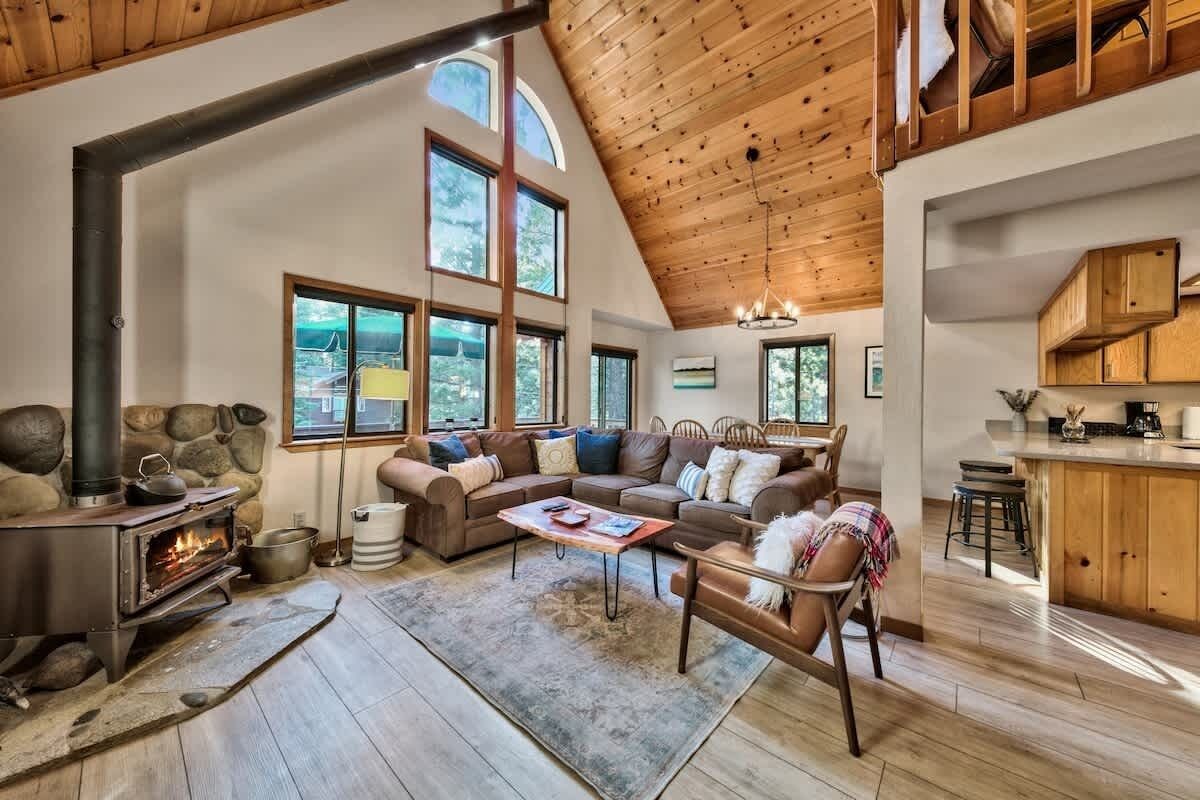 A cozy living room with a fireplace, large windows, a stone wall, and a high ceiling.