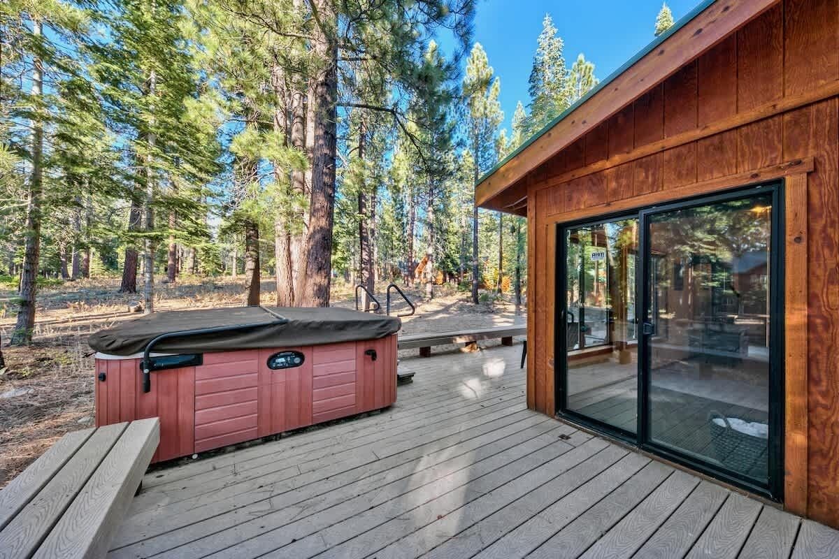 Terraza de madera con jacuzzi, puerta de vidrio que da a un área cubierta y un bosque al fondo.