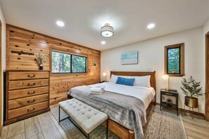 A bedroom with a wooden wall, a bed, a bench, a dresser, and a nightstand with a lamp.
