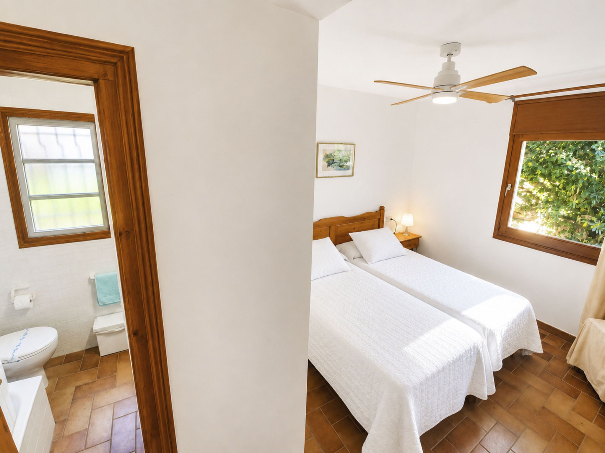 A bedroom with two beds, a ceiling fan, a window with a view of greenery, and a small bedside table with a lamp.