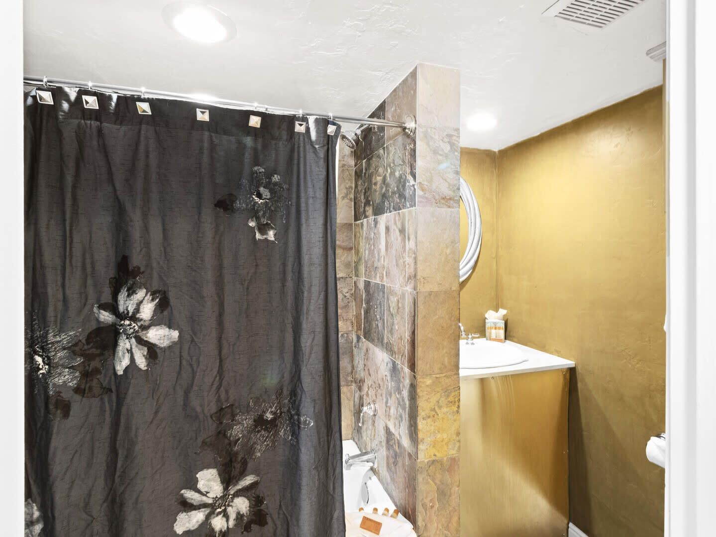 Casa de banho com cortina de chuveiro estampada com motivos florais, parede revestida a mármore, parede com detalhes dourados e lavatório com espelho.