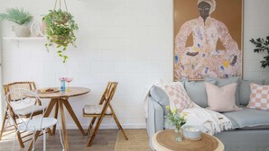 A cozy living room with a round wooden table, two wicker chairs, a grey sofa with patterned cushions, and a hanging plant. - Palm Springs Oasis, short walk to Shelly Beach (Bateau Bay)