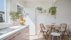 A kitchen with a white countertop, pink cabinets, and a window. There is a dining area with a round table, two chairs, and hanging plants. - Palm Springs Oasis, short walk to Shelly Beach (Bateau Bay)